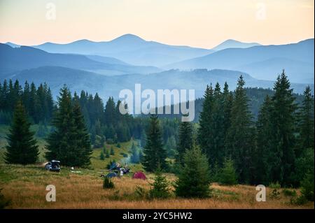 Campingplatz aus der Vogelperspektive in den Bergen, mit mehreren bunten Zelten auf einer grasbewachsenen Rodung. Camper entspannen unter klarem blauen Himmel, umgeben von hohen immergrünen und Panoramablick auf entfernte Gipfel. Stockfoto