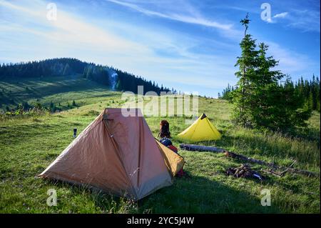 Zwei Zelte, orange und gelb, auf einem grasbewachsenen Hügel unter klarem blauem Himmel. Campingausrüstung in der Nähe verstreut, ferne Rauchwolke steigt aus dem Feuer auf. Umgeben von sanften Hügeln und dichten Kiefern. Stockfoto