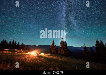 Silhouetten von Touristen, die auf dem Campingplatz am Lagerfeuer ruhen, mit leuchtenden Zelten in der Nähe unter Sternenhimmel. Die Milchstraße erstreckt sich über die Berge und wirft ein heiteres und magisches Licht über die beschauliche Berglandschaft. Stockfoto