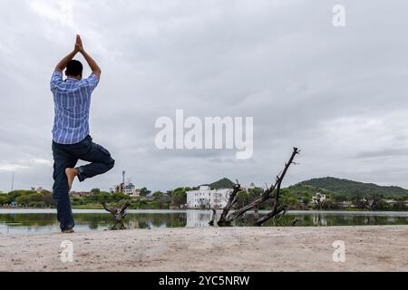 Junger Mann, der meditiert und Yoga in der Nähe eines unberührten ruhigen Sees mit dramatischem Himmel am Morgen macht Stockfoto