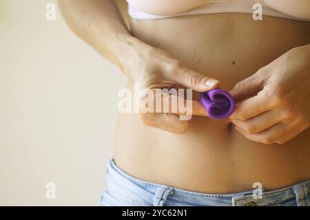 Junge Frau in BH und Jeans, die einen kleinen gefalteten Menstruationsbecher hält. Gesündere und bequemere Alternative zu Tampons. Verwendung wiederverwendbarer Menstruationsbecher Stockfoto