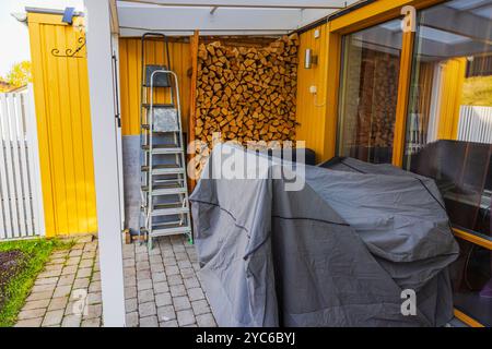 Holzstapel, überdachte Möbel und Leiter im gelben Haus im Hinterhof mit Terrasse. Schweden. Stockfoto