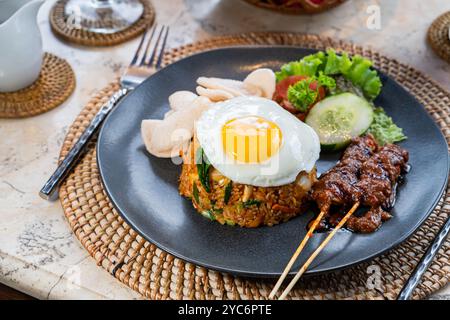 Ein Teller mit gebratenem Reis, garniert mit einem sonnigen Ei, serviert mit Gurkenscheiben, Salat und Garnelencrackern, dazu Satay-Spieße mit Pean Stockfoto
