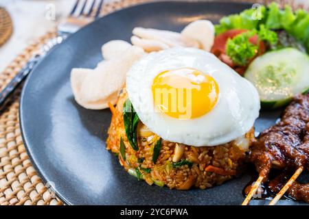 Ein Teller mit gebratenem Reis, garniert mit einem sonnigen Ei, serviert mit Gurkenscheiben, Salat und Garnelencrackern, dazu Satay-Spieße mit Pean Stockfoto