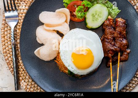 Ein Teller mit gebratenem Reis, garniert mit einem sonnigen Ei, serviert mit Gurkenscheiben, Salat und Garnelencrackern, dazu Satay-Spieße mit Pean Stockfoto
