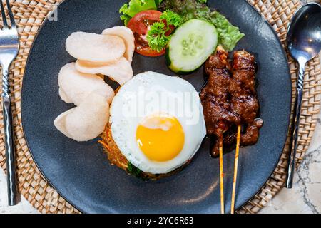 Ein Teller mit gebratenem Reis, garniert mit einem sonnigen Ei, serviert mit Gurkenscheiben, Salat und Garnelencrackern, dazu Satay-Spieße mit Pean Stockfoto