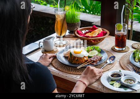 Ein Teller mit gebratenem Reis, garniert mit einem sonnigen Ei, serviert mit Gurkenscheiben, Salat und Garnelencrackern, dazu Satay-Spieße mit Pean Stockfoto