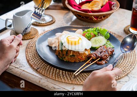 Ein Teller mit gebratenem Reis, garniert mit einem sonnigen Ei, serviert mit Gurkenscheiben, Salat und Garnelencrackern, dazu Satay-Spieße mit Pean Stockfoto
