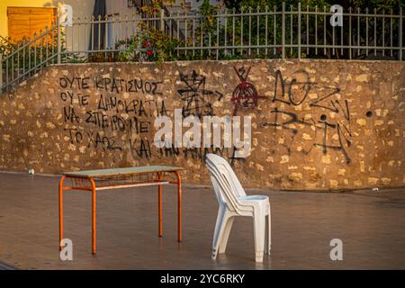 Eine Szene auf Kreta mit einem einzelnen weißen Plastikstuhl und einem Metalltisch vor einer Wand mit Graffiti. Der Text und die Symbole an der Wand verleihen dem Bild einen Hauch Stockfoto