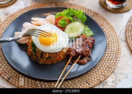 Ein Teller mit gebratenem Reis, garniert mit einem sonnigen Ei, serviert mit Gurkenscheiben, Salat und Garnelencrackern, dazu Satay-Spieße mit Pean Stockfoto