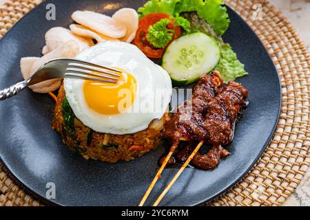 Ein Teller mit gebratenem Reis, garniert mit einem sonnigen Ei, serviert mit Gurkenscheiben, Salat und Garnelencrackern, dazu Satay-Spieße mit Pean Stockfoto
