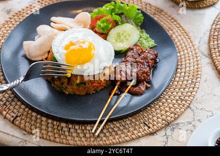 Ein Teller mit gebratenem Reis, garniert mit einem sonnigen Ei, serviert mit Gurkenscheiben, Salat und Garnelencrackern, dazu Satay-Spieße mit Pean Stockfoto