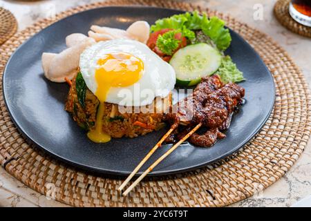 Ein Teller mit gebratenem Reis, garniert mit einem sonnigen Ei, begleitet von Satay-Spießen, Gurkenscheiben, Salat und Tomaten. Das Gericht wird auf einem bl serviert Stockfoto