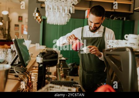 In einem warmen und einladenden Café gießt ein engagierter Barista fachmännisch Milch in eine Kaffeetasse und sorgt so für ein angenehmes Erlebnis für Gäste. Stockfoto
