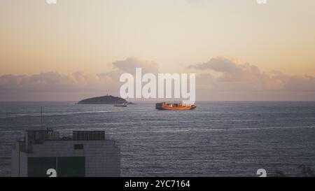 Frachtschiff, das während des Sonnenuntergangs auf einem ruhigen Meer segelt, beleuchtet von warmem, goldenem Licht. Im Hintergrund ist eine Insel mit einem Leuchtturm sichtbar, gerahmt b Stockfoto