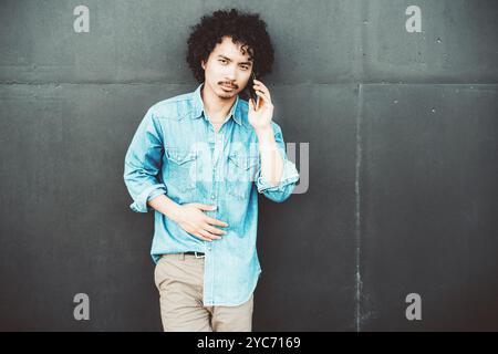 Ein junger Mann mit lockigen Haaren in einem lässigen blauen Jeanshemd steht vor einer dunklen Wand und hält ein Smartphone am Ohr. Er scheint konzentriert und engagiert zu sein Stockfoto