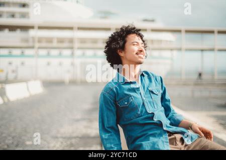 Ein Mann mit lockigen Haaren in einem blauen Jeanshemd genießt einen sonnigen Tag und lächelt, während er draußen sitzt. Der Hintergrund zeigt ein verschwommenes Schiff und eine Uferpromenade, die eine ergibt Stockfoto