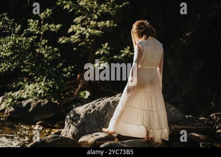 Frau in weißem Kleid, die auf Felsen an einem ruhigen Bach in einem sonnendurchfluteten Wald steht Stockfoto