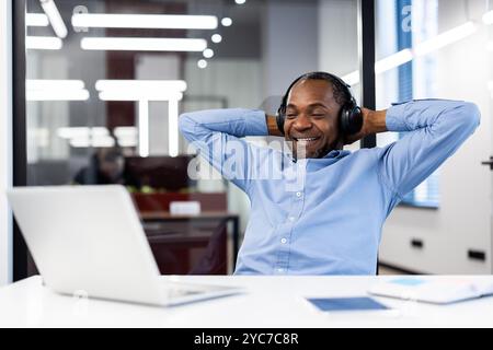 Reifer afroamerikanischer Geschäftsmann in blauem Hemd entspannt sich im Büro und hört Musik mit Kopfhörern. Heller, moderner Arbeitsbereich sorgt für beruhigende Atmosphäre Stockfoto