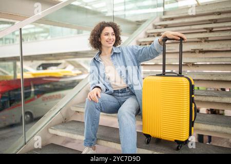 Die junge Frau ruht auf einer Treppe mit gelbem Koffer an einem geschäftigen Reiseterminal Stockfoto