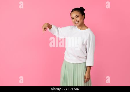 Die junge Frau zeigt nach unten, während sie vor einem leuchtenden rosa Hintergrund lächelt Stockfoto