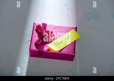 Ein Lichtstrahl fällt auf eine pinkfarbene Geschenkbox mit einem Schreibpapier und einer handgeschriebenen Inschrift Danke. Kopierbereich. Stockfoto