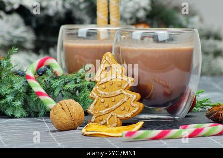 Zwei Klarglasbecher gefüllt mit heißem Kakao, ein Lebkuchenkeks in Form eines Weihnachtsbaums, ein Tannenzweig, Nüsse und ein Zuckerrohr ergänzen das Stockfoto