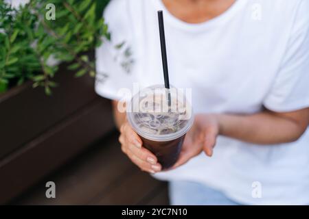Nahaufnahme einer unerkennbaren Frau in weißem T-Shirt, die eine Plastiktasse zum Mitnehmen mit kaltem Kaffeetrink im Freien hält. Stockfoto