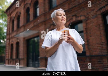 Eine hübsche ältere Frau in lässigen Kleidern, die draußen Eiskaffee trinkt, sieht nachdenklich aus in urbaner Umgebung mit Ziegelbauten auf verschwommenem Hintergrund. Stockfoto