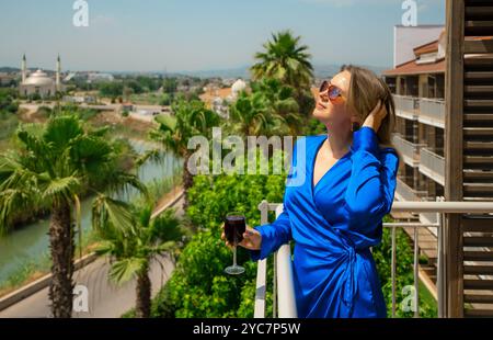Frau in blauem Kleid mit Rotwein genießt Urlaub auf dem Balkon. Stockfoto