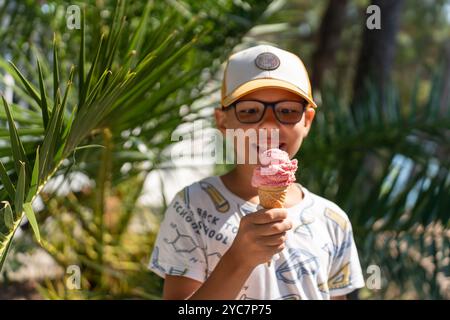 Glücklicher Junge mit Brille und Mütze, der an einem hellen Sommertag eine Erdbeereiskrem hält. Stockfoto