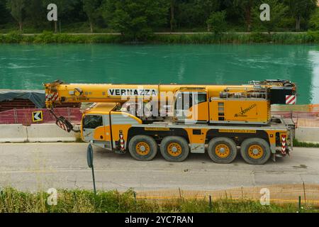 Peschiera del Garda, Italien - 13. Juni 2023: Ein großer gelber Kran steht am Straßenrand neben einem lebhaften türkisfarbenen Fluss und zeigt sein funktionales Design Stockfoto