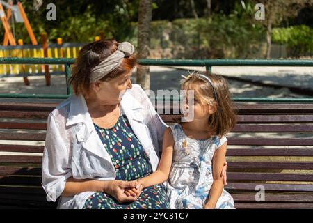 Seniorin und niedliches Mädchen sitzen auf der Parkbank, blicken sich mit einem warmen Lächeln an und teilen sich zärtliche und liebevolle Momente an sonnigen Tagen Stockfoto