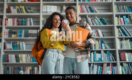 Zwei Studenten Klassenkameraden Afroamerikaner Mann Inder Latino Arabische Frau Handy Smartphone Bibliothek Lächeln Vergnügen Lachen Kommunikation Internet Stockfoto