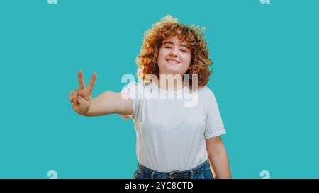 Siegesgeste. Fröhliches, lockiges Mädchen Lächeln zeigt ein Friedenszeichen, das eine sorglose und positive Einstellung auf blauem Kopierraum-Hintergrund widerspiegelt. Stockfoto