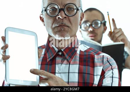 Zwei nervöse Wissenschaftler mit dicker Brille halten und zeigen auf ein digitales Tablet mit leerem Bildschirm, während einer von ihnen Notizen zu einem Buch macht Stockfoto