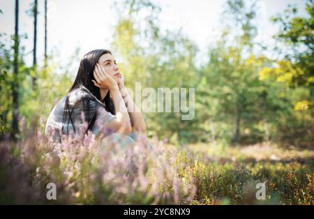 Die junge Frau ist umgeben von blühenden Wildblumen und grünen Bäumen, die in Gedanken verloren sind, während sie sich an einem ruhigen Nachmittag im warmen Sonnenlicht sonnt. Stockfoto