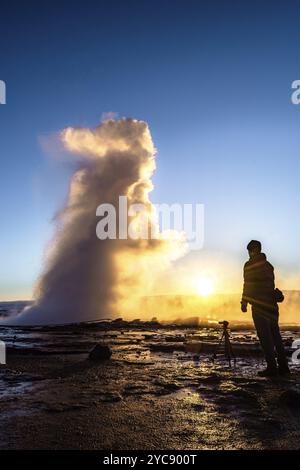 Stehende Person fotografiert einen ausbrechenden Geysir bei Sonnenuntergang in Island, Strokkur Stockfoto