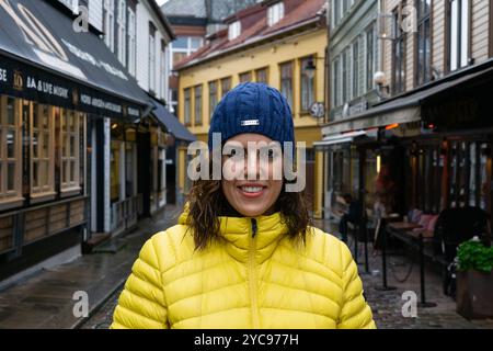 Eine junge Brünette, etwa 30 Jahre alt, trägt eine gelbe Pufferjacke und einen blauen Strickmütze, lächelt direkt in die Kamera, während sie auf einem rai steht Stockfoto
