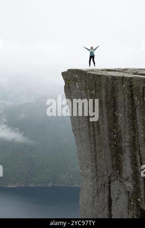 Eine Frau, die an einem regnerischen Tag im Lysefjord in Norwegen am Rande der Preikestolen-Klippe, auch bekannt als Pulpit Rock, vor Freude springt. Norwegen Tourismuskonsum Stockfoto