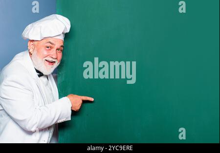 Leitender Chefkoch. Portrait des Chefkochs mit Handzeichen auf Tafel Hintergrund. Aufgeregt Mann Koch Koch trägt Uniform zeigt Kopie Raum Text. Stockfoto