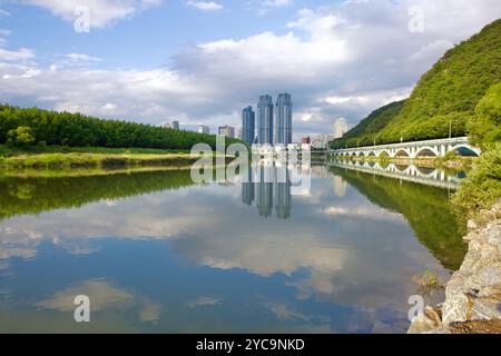 Ulsan, Südkorea - 9. Juli 2019: Ruhiges Wasser des Taehwa River, das die Wolken reflektiert, mit dem Taehwa River National Garden auf der linken Uferseite und einem Fahrrad Stockfoto