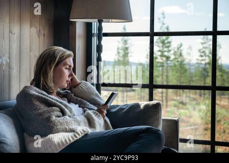 Die Frau sitzt bequem auf einer Couch in einer Hütte, vertieft in ihr E-Reader Buch. Sonnenlicht strömt durch ein großes Fenster und offenbart eine Waldlandschaft Stockfoto