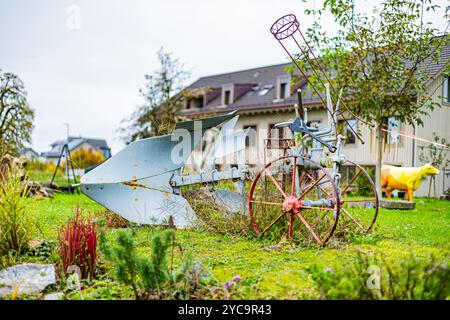 Ein rostiger alter Pflug sitzt auf einem Hof neben einem Haus. Der Hof ist mit verschiedenen Pflanzen und Blumen gefüllt, darunter einige rote Blumen. Die Szene ist irgendwie Stockfoto