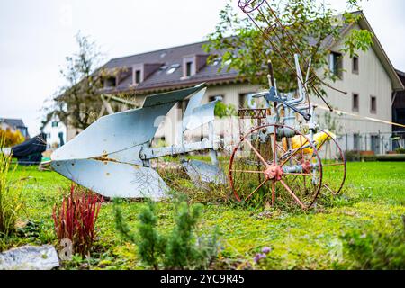 Ein rostiger alter Pflug sitzt auf einem Hof neben einem Haus. Der Hof ist mit verschiedenen Pflanzen und Blumen gefüllt, darunter einige rote Blumen. Die Szene ist irgendwie Stockfoto