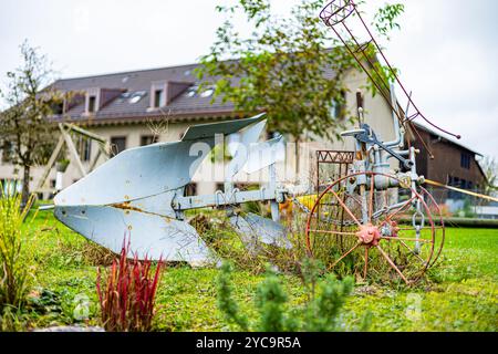 Ein rostiger alter Pflug sitzt auf einem Hof neben einem Haus. Der Hof ist mit verschiedenen Pflanzen und Blumen gefüllt, darunter einige rote Blumen. Die Szene ist irgendwie Stockfoto