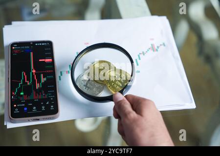 Junger indischer Kryptohändler oder -Broker, der Bitcoin mit einer Lupe auf dem Finanzchart überprüft. Mobile Anzeige von Marktdaten. Stockfoto