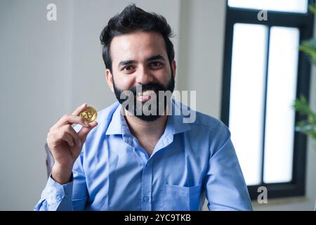 Fröhlicher junger indischer Geschäftsmann, der Bitcoin, Kryptowährung, in der Hand hält, während er in die Kamera schaut. Investitions- und Finanzplanungskonzept. Stockfoto