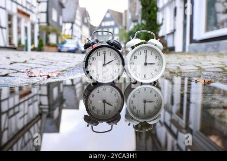 Zwei Wecker stehen in der Altstadt von Freudenberg in einer Pfuetze Pfütze und spiegeln sich darin. Ein Wecker zeigt drei Uhr, der andere zwei Uhr. Die Uhren werden auf Winterzeit umgestellt. In der Nacht, am 27.10.2024 werden die Uhren von 03:00 Uhr auf 02,00 Uhr zurueckgestellt zurückgestellt. Symbolbild zum Thema, Winterzeit, Zeitumstellung, Uhrenumstellung am 22.10.2024 in Freudenberg/Deutschland. *** Zwei Wecker stehen in einer Wasserpfütze in der Altstadt von Freudenberg und spiegeln sich darin wider ein Wecker zeigt drei Uhr, die anderen zwei Uhr werden in Winte geändert Stockfoto