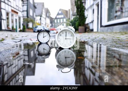 Zwei Wecker stehen in der Altstadt von Freudenberg in einer Pfuetze Pfütze und spiegeln sich darin. Ein Wecker zeigt drei Uhr, der andere zwei Uhr. Die Uhren werden auf Winterzeit umgestellt. In der Nacht, am 27.10.2024 werden die Uhren von 03:00 Uhr auf 02,00 Uhr zurueckgestellt zurückgestellt. Symbolbild zum Thema, Winterzeit, Zeitumstellung, Uhrenumstellung am 22.10.2024 in Freudenberg/Deutschland. *** Zwei Wecker stehen in einer Wasserpfütze in der Altstadt von Freudenberg und spiegeln sich darin wider ein Wecker zeigt drei Uhr, die anderen zwei Uhr werden in Winte geändert Stockfoto